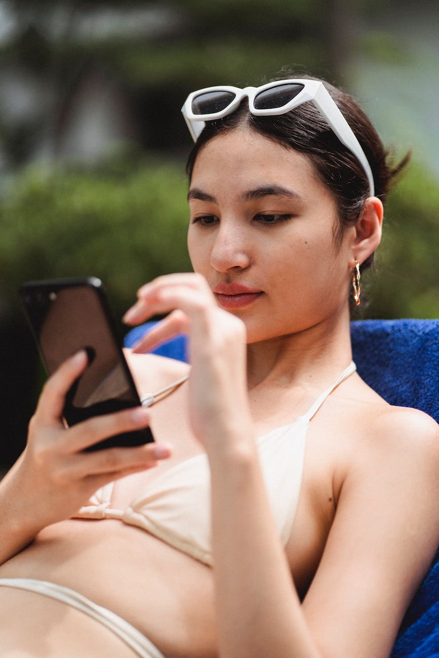 focused young ethnic lady in swimwear surfing smartphone lying on sunbed